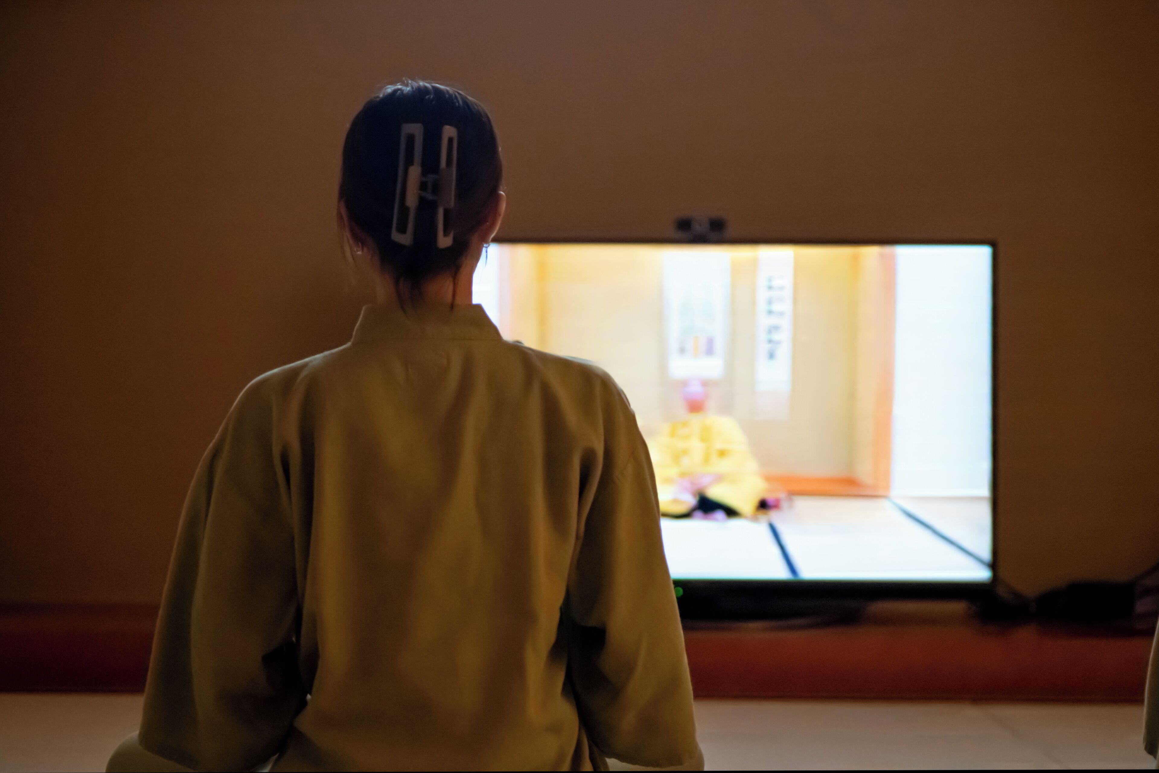 Eine Person sitzt vor einem Fernseher in einem Zimmer mit einer schlichten Wand und einem Fenster.