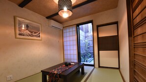 Ein traditioneller japanischer Raum mit Tatami-Bodenbelag, einem niedrigen Tisch mit gemustertem Tischtuch, einer Shoji-Schiebetür und einem gerahmten Kunstwerk an der Wand.
