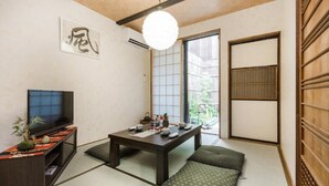 Ein traditioneller japanischer Raum mit Tatami-Böden, einem niedrigen Tisch mit Kissen, einem Fernseher auf einem Regal und einer Schiebetür, die zu einem Garten führt.