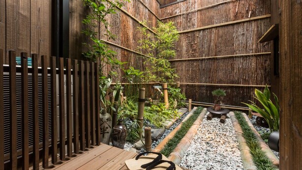 Eine Holzterrasse mit einem kleinen Tisch und Stühlen, umgeben von einem Bambuszaun und verschiedenen Pflanzen.