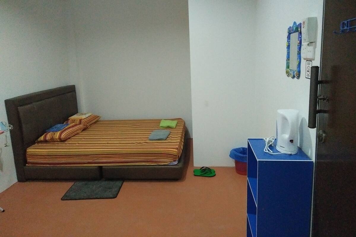Pequeña habitación con cama individual, cabecera de madera, ropa de cama a rayas, un tapete verde, una cafetera blanca y un mueble azul.