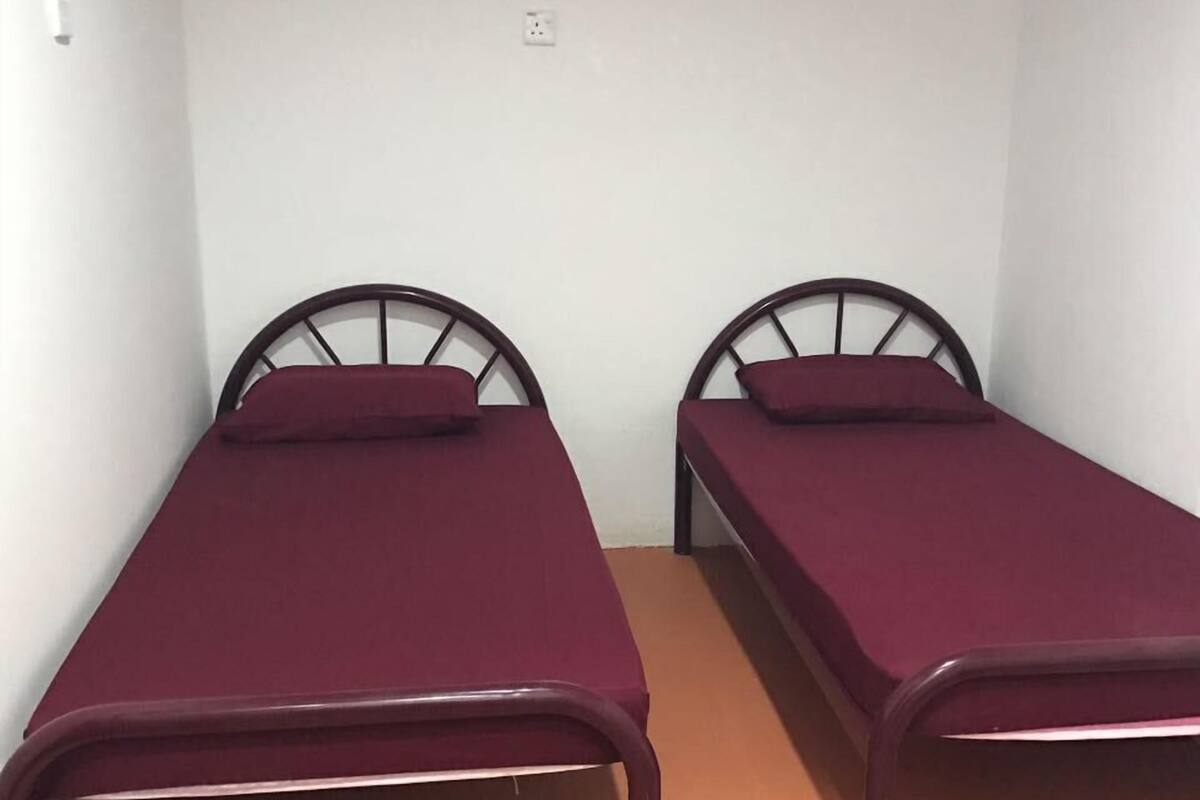Dos camas individuales con estructura metálica y ropa de cama roja en una habitación pequeña con paredes blancas lisas y suelo naranja.