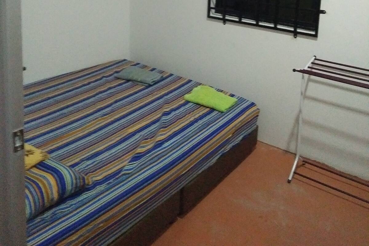 Una habitación pequeña con una cama individual, colcha a rayas, ventana con rejas y un tendedero en la pared.