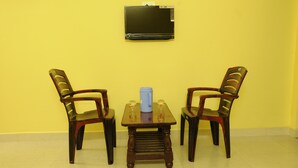 Ein Zimmer mit gelber Wand, einem Holztisch mit zwei Stühlen, einer blauen Tasse und einem an der Wand montierten Fernseher.