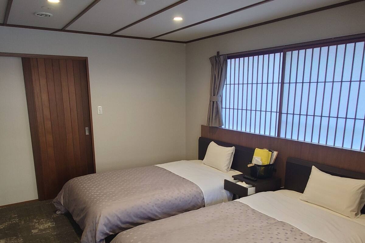 Une chambre d’hôtel avec deux lits, une porte en bois, une fenêtre avec des barreaux et un plafond doté d’un éclairage encastré.