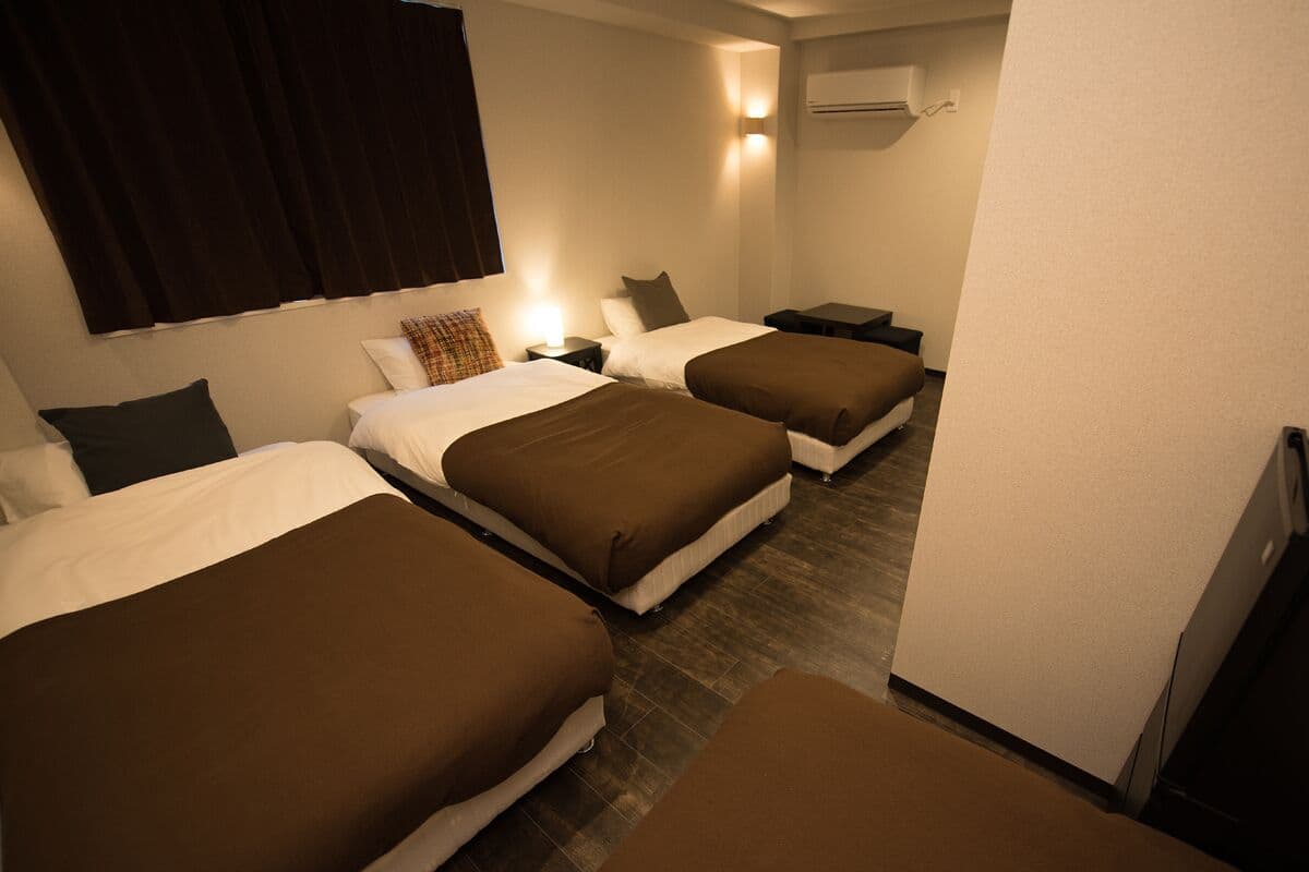 A hotel room with three beds, each with a brown bedspread, white pillows, and a small bedside table with a lamp.