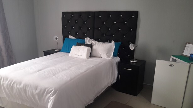 A neatly made bed with white bedding and a black headboard with diamond patterns. There are two bedside tables, one with a lamp and the other with a small appliance. A brown doormat is placed in front of the bed. - Jessma Bed and Breakfast (Walvis Bay)