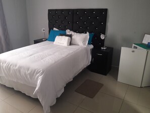 A neatly made bed with white bedding and a black headboard with diamond patterns. There are two bedside tables, one with a lamp and the other with a small appliance. A brown doormat is placed in front of the bed.