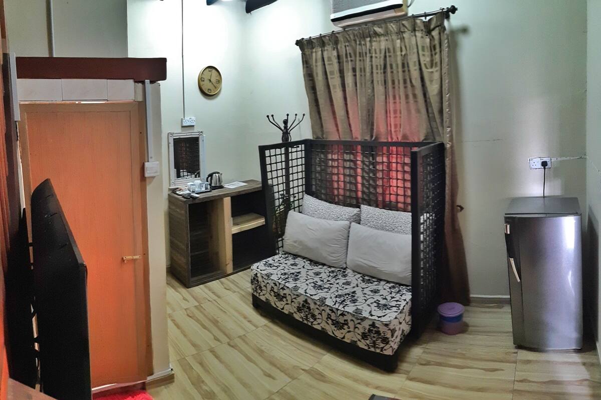 A compact room with a bed, a small desk, a refrigerator, and a clock on the wall.