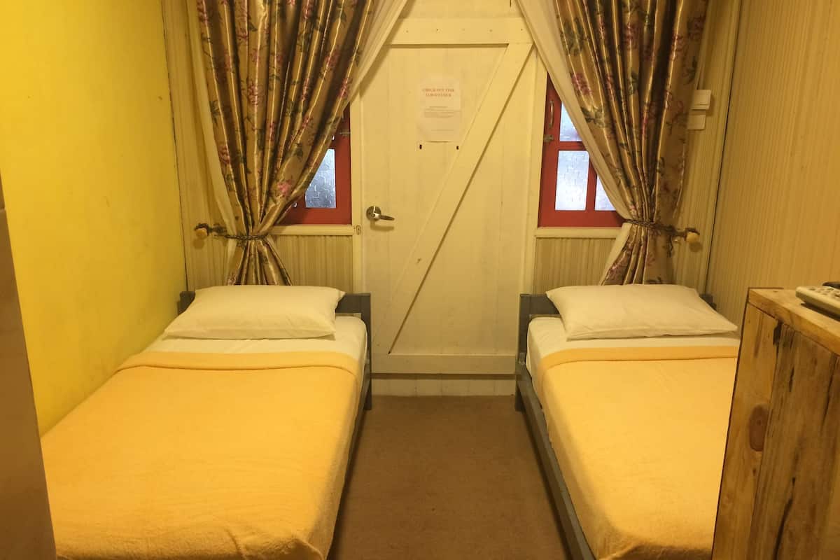 A small, narrow room with two single beds, a wooden wardrobe, and a door with a check-out time sign.