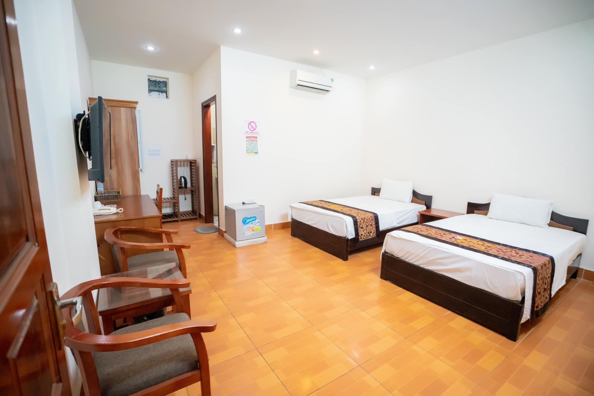 A hotel room with two beds, a desk, chairs, an air conditioner, and a refrigerator.