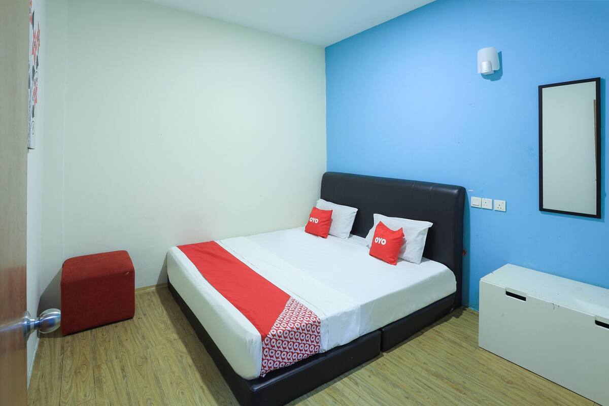A bedroom with a bed, a red ottoman, a white chest of drawers, and a blue wall.