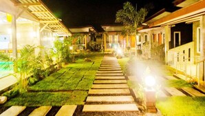 Vista noturna de um corredor de hotel com um caminho central, rodeado por quartos bem iluminados e vegetação exuberante.