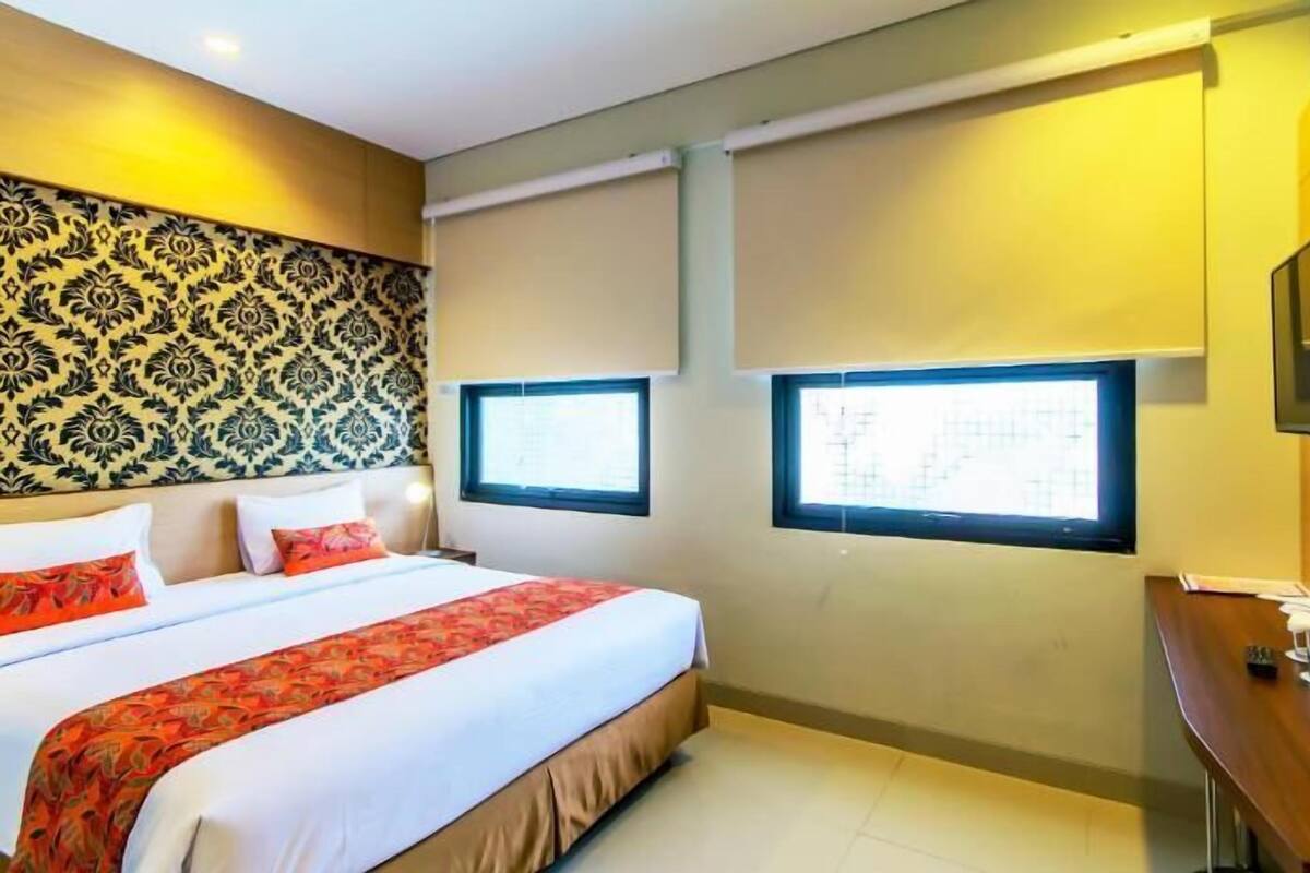 A hotel room with a large bed, two windows, a TV, and a desk.