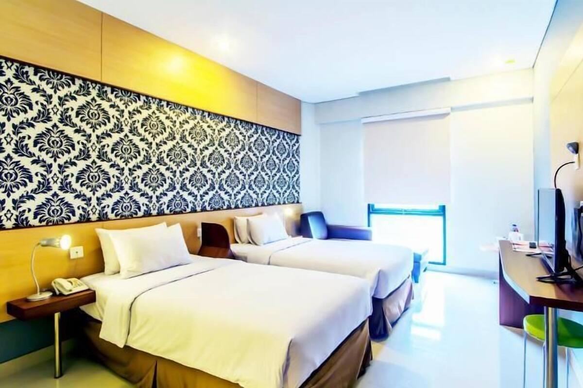 A hotel room with two beds, a large patterned wall, a desk with a computer, and a window with blinds.