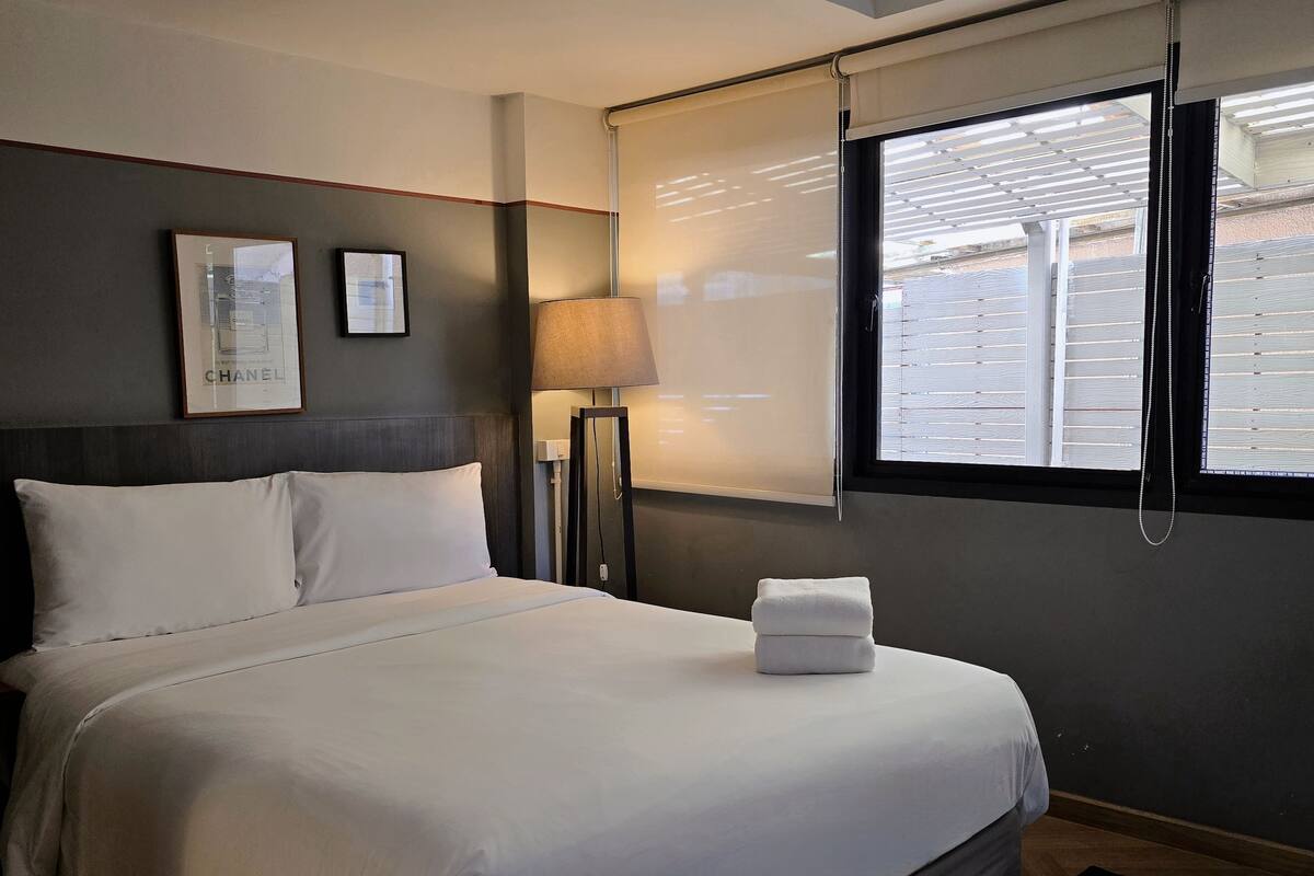 A neatly made bed with white linens, a bedside lamp, a framed picture on the wall, and a window with blinds.