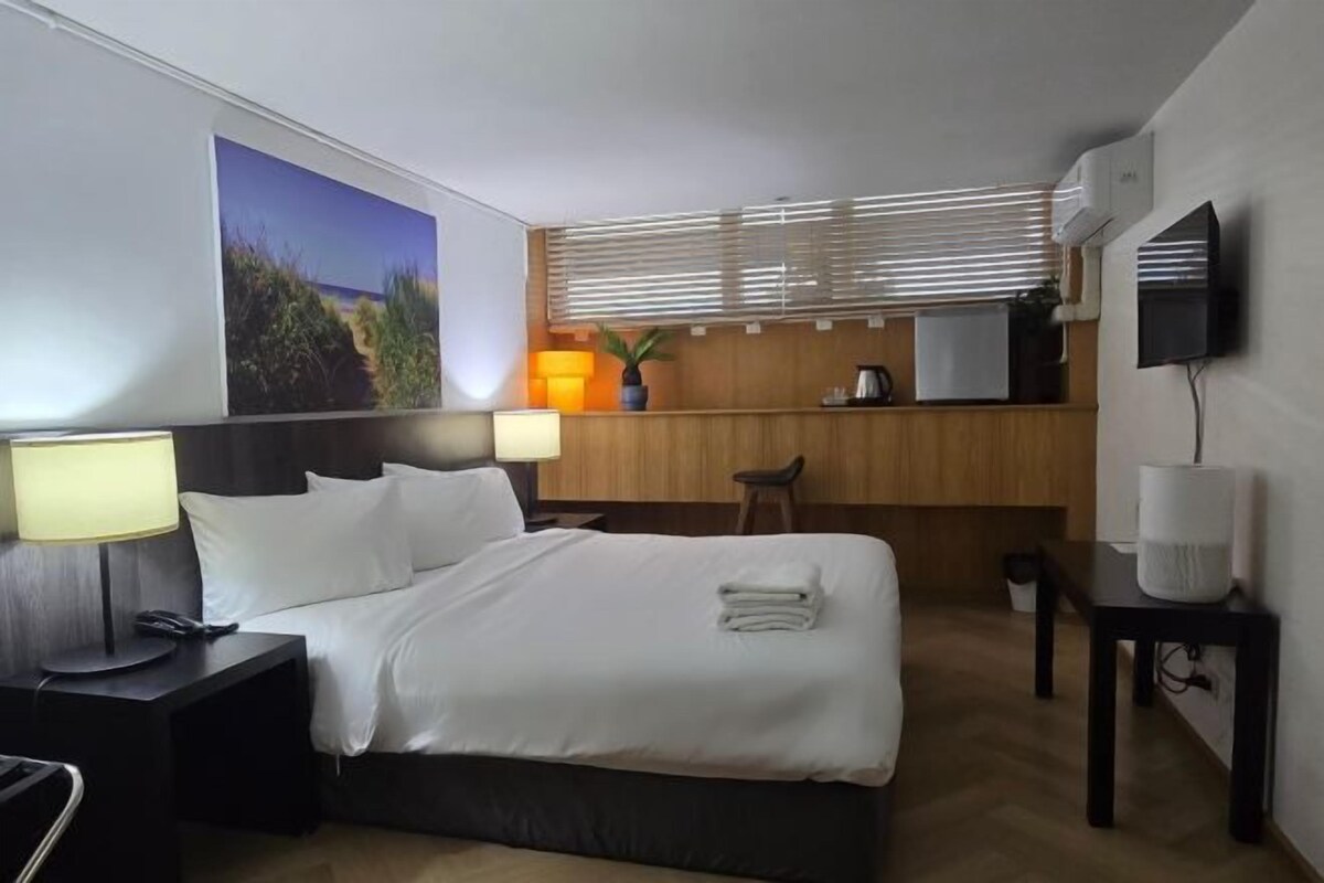 A hotel room with a bed, bedside table, lamp, and a view of a beach through the window.