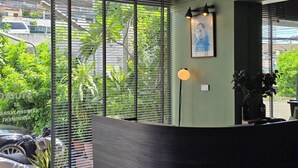 A modern reception area with a curved desk, large windows with blinds, a framed portrait, and a floor lamp. - Somewhere to Escape - Hometel (Bangkok)