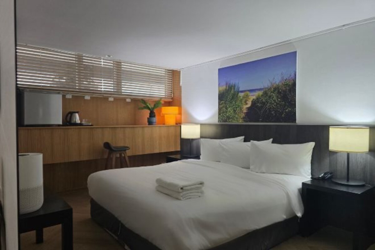 A hotel room with a bed, bedside lamps, a desk with a lamp, a chair, a TV, and a painting on the wall.