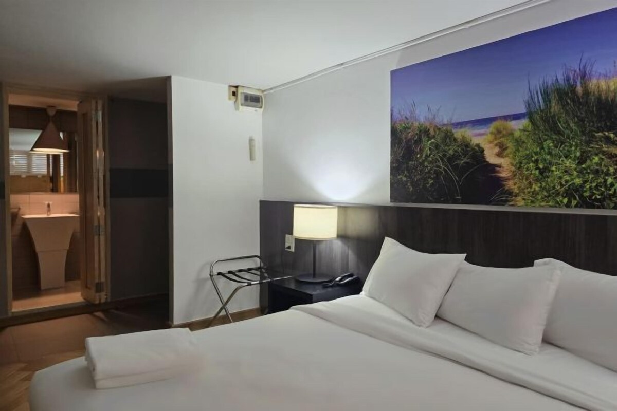 A hotel room with a bed, a nightstand with a lamp, a painting of a beach scene, and a bathroom visible through an open door.