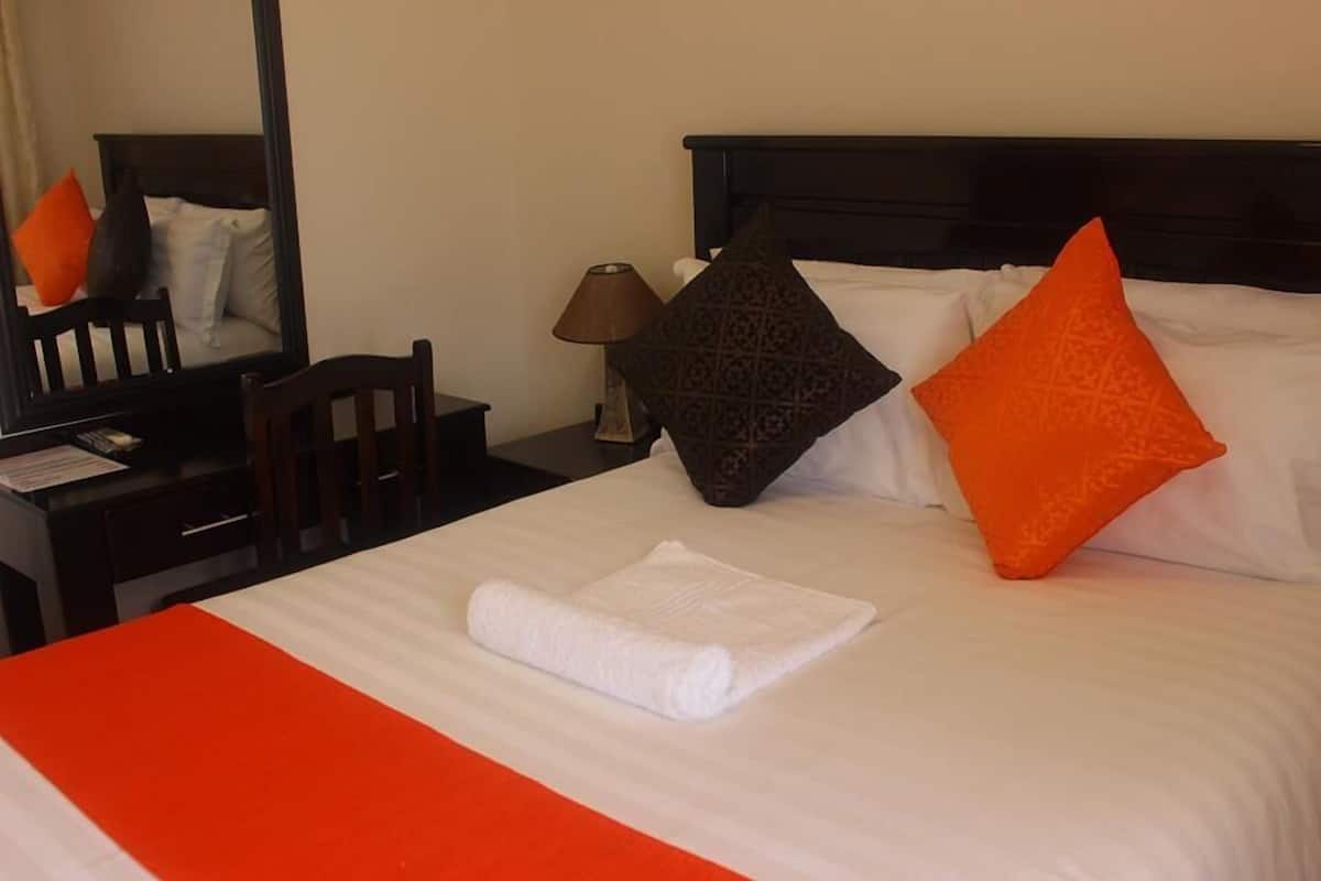 A neatly made bed with a white and orange bedspread, a folded white towel, and two pillows, one orange and one patterned.