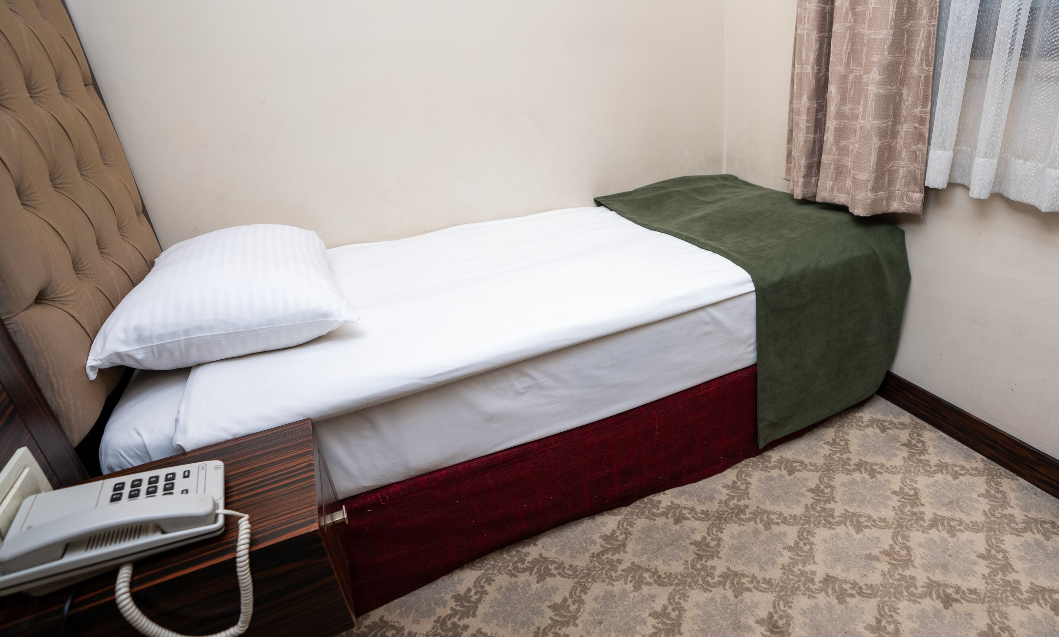 Ein kleines Zimmer mit einem Einzelbett, einem Telefon, einer grünen Bettdecke und einem gemusterten Teppich.