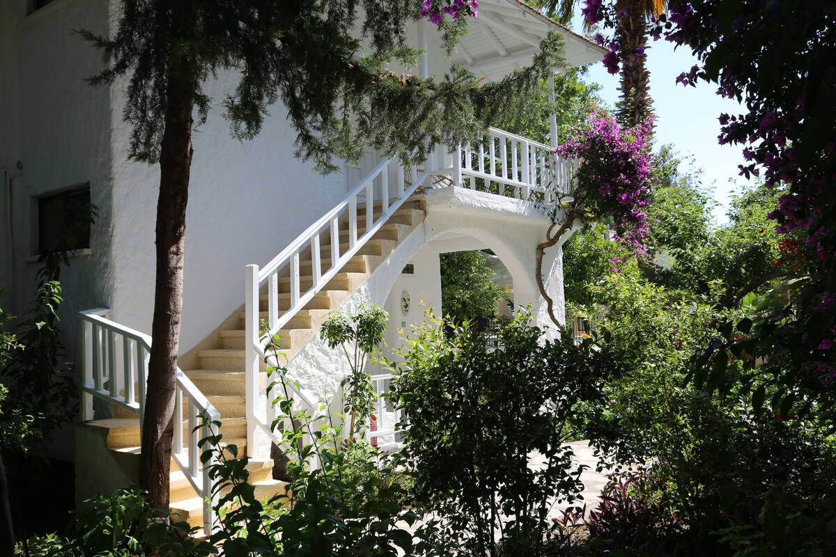 Un bâtiment blanc avec un escalier menant à un balcon, entouré d’une végétation luxuriante et de plantes en fleurs.