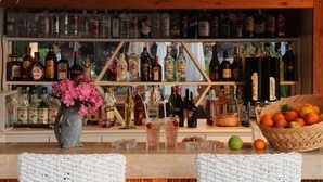 Eine Bar mit einem Reetdach, Flaschenregalen, einer Vase mit rosa Blumen und Korbstühlen.