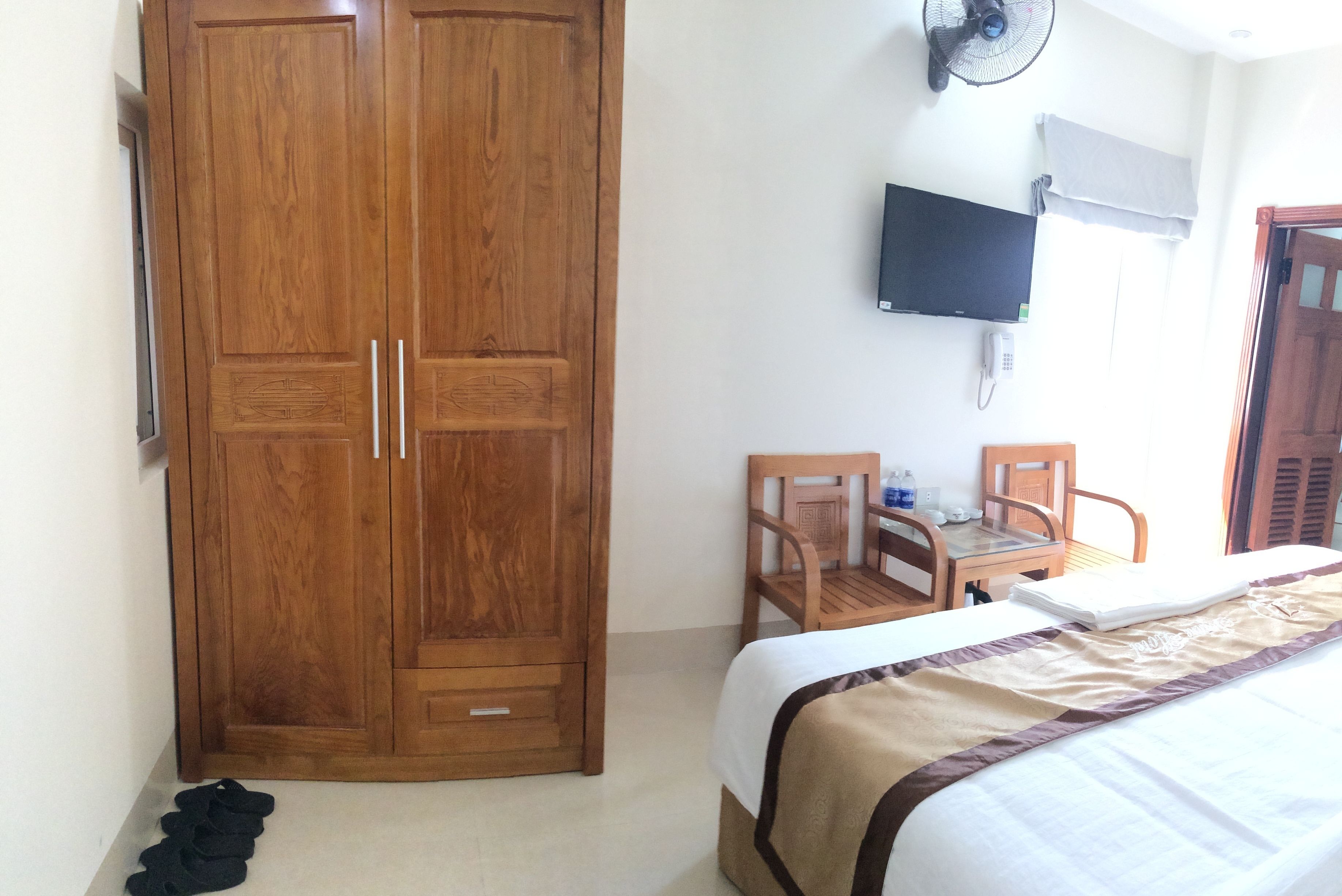 Une chambre avec une armoire en bois, un lit recouvert d’une couverture beige et marron, un bureau en bois avec une chaise, un téléviseur fixé au mur et un ventilateur de plafond.