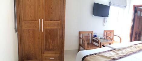 Une chambre avec une armoire en bois, un lit recouvert d’une couverture beige et marron, un bureau en bois avec une chaise, un téléviseur fixé au mur et un ventilateur de plafond.