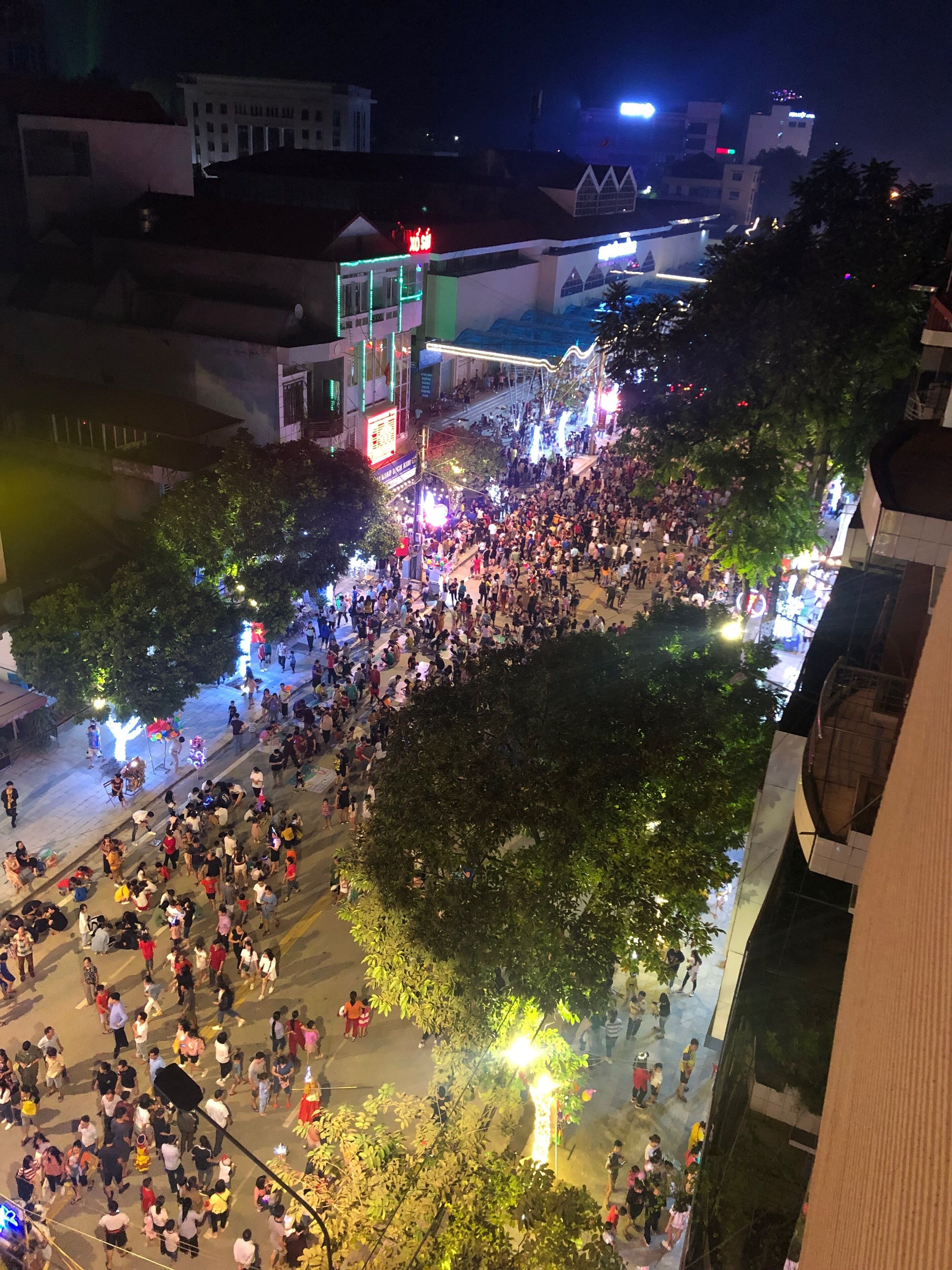 A crowded street scene at night with illuminated buildings and trees.