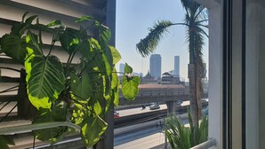 A view from a window with a balcony, featuring a cityscape with tall buildings and a bridge, alongside some greenery. - Sathorn Terrace Apartment (Bangkok)