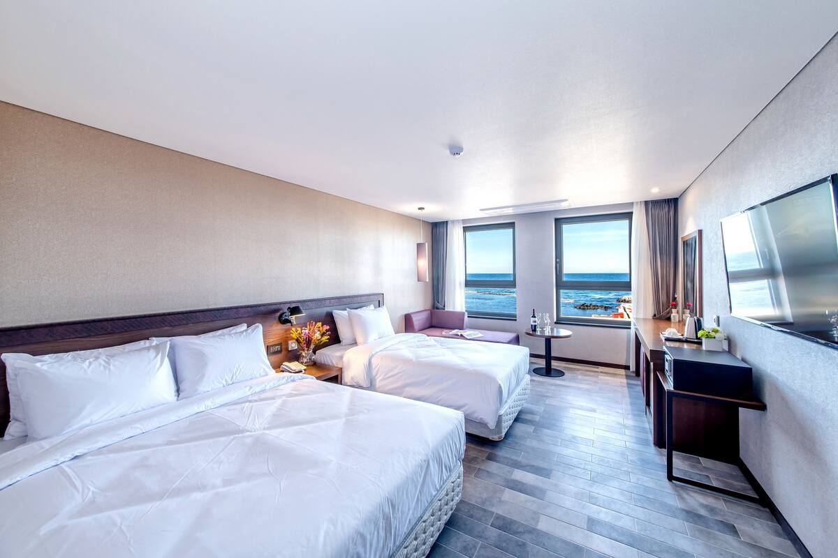 A hotel room with two beds, a small seating area, a desk, and a large window with a view of the sea.