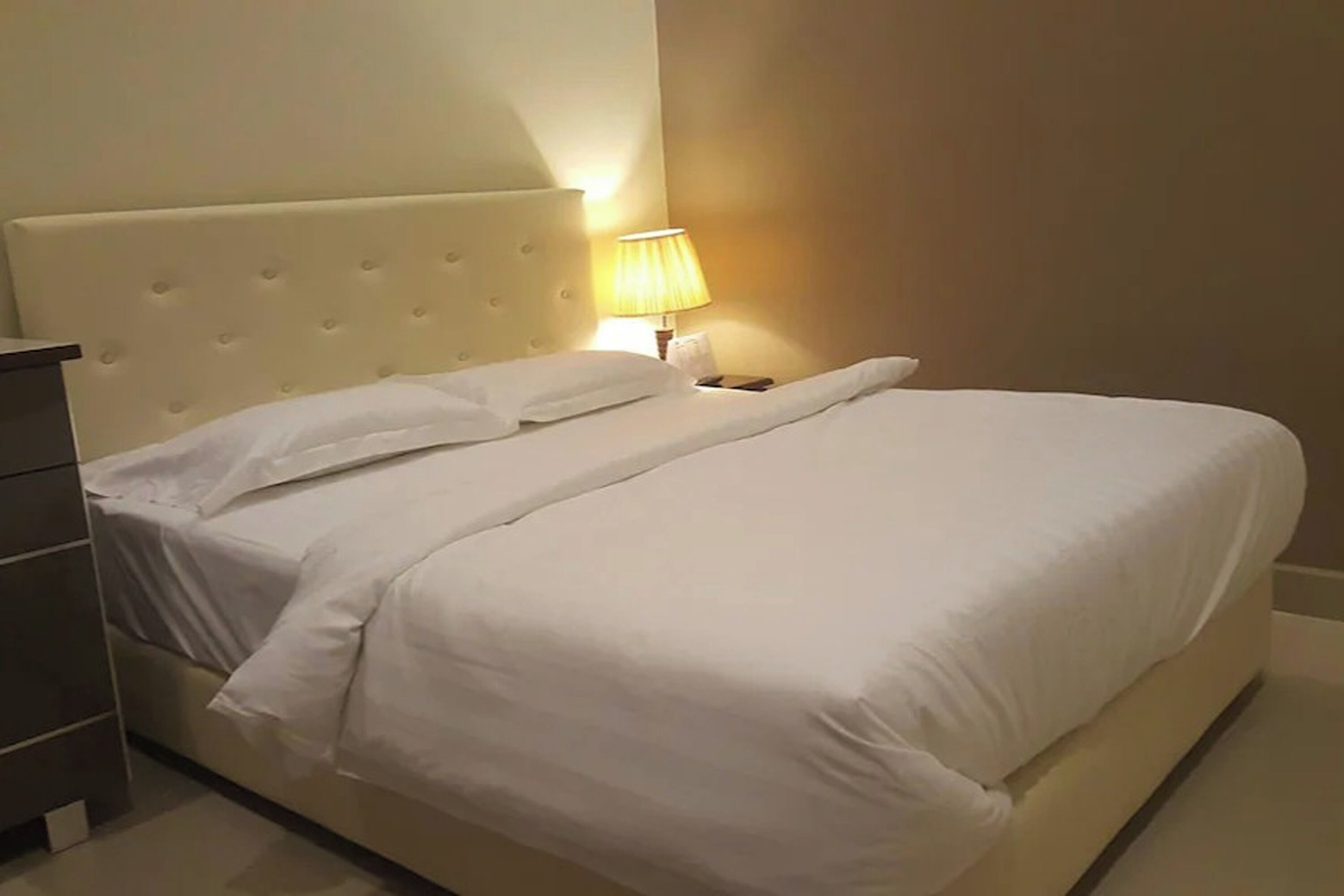 A neatly made bed with white linens, a tufted headboard, and a bedside lamp.