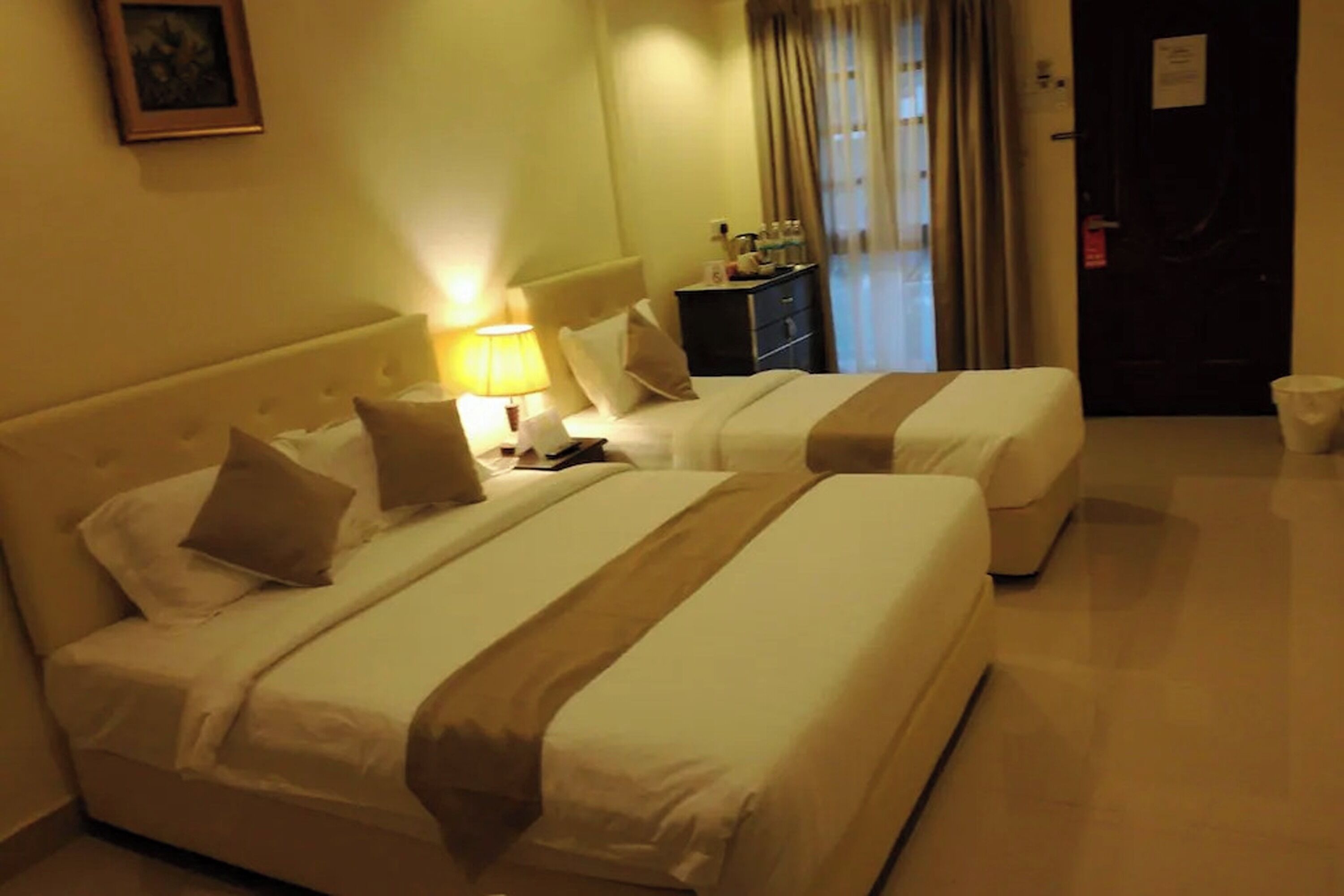 A hotel room with two beds, a nightstand, a lamp, a dresser, and a door.