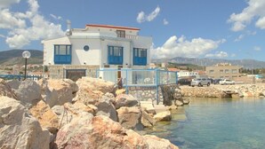 A coastal building with a white facade and blue accents, situated near a rocky shoreline with clear water. - Uzum Iskelesi Butik Otel (Karaburun)
