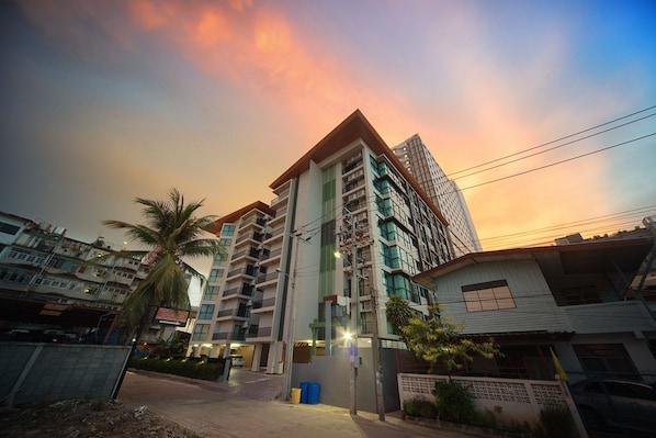 A multi-story residential building with balconies, a street in front, and a sunset sky.