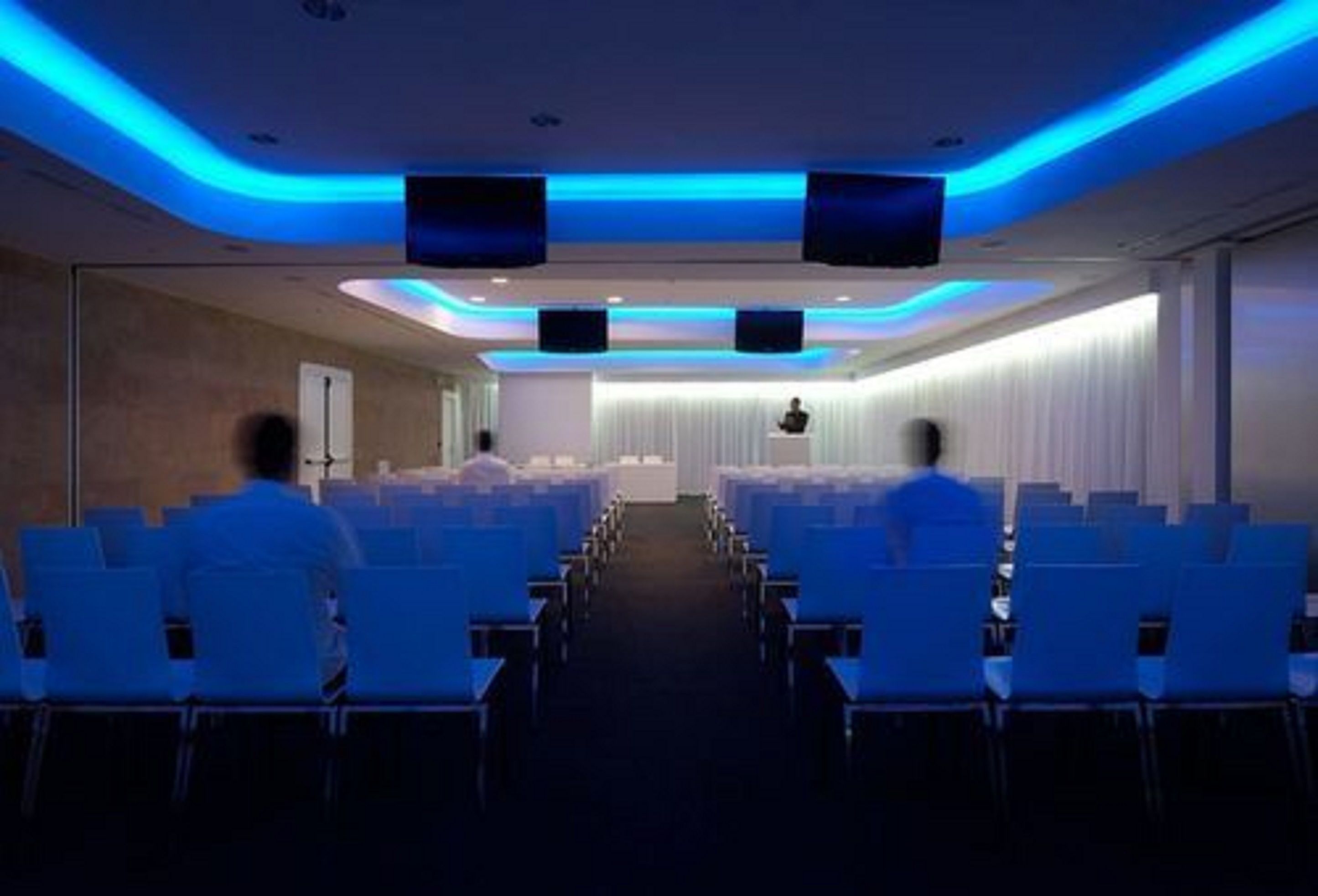 A conference room with rows of white chairs, a stage at the front, and blue ambient lighting.
