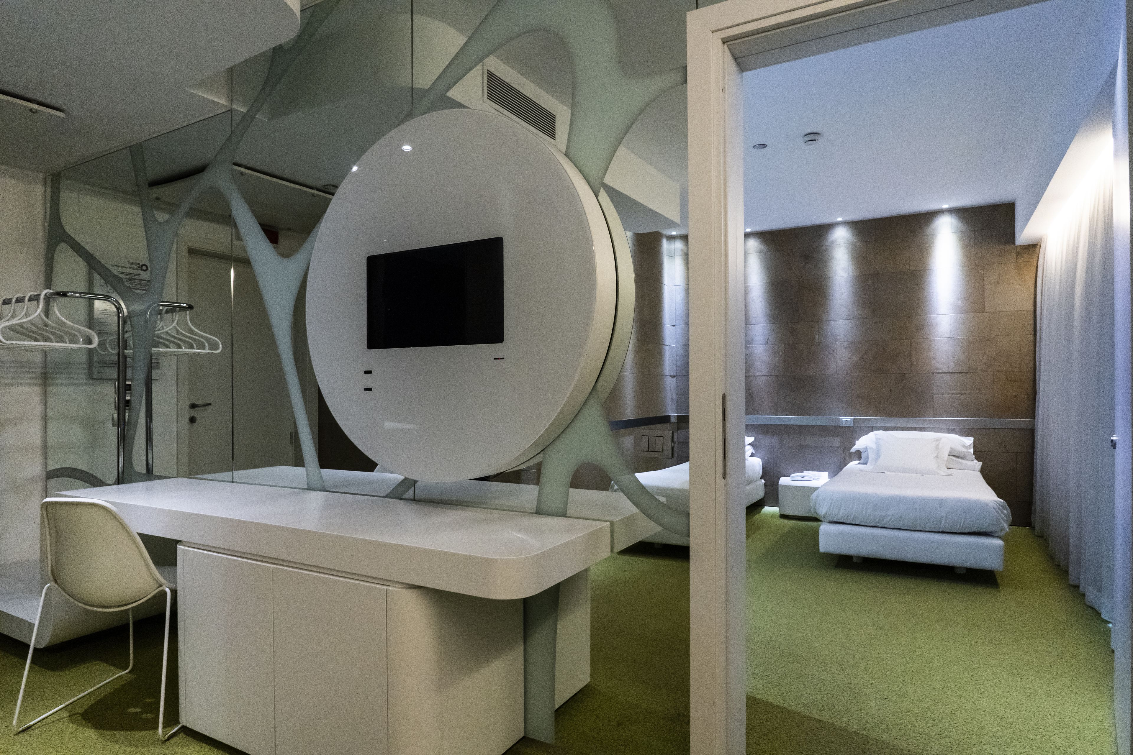 Et moderne hotelværelse med et stort spejl, et fladskærms-TV, en seng og et skrivebord med stol.