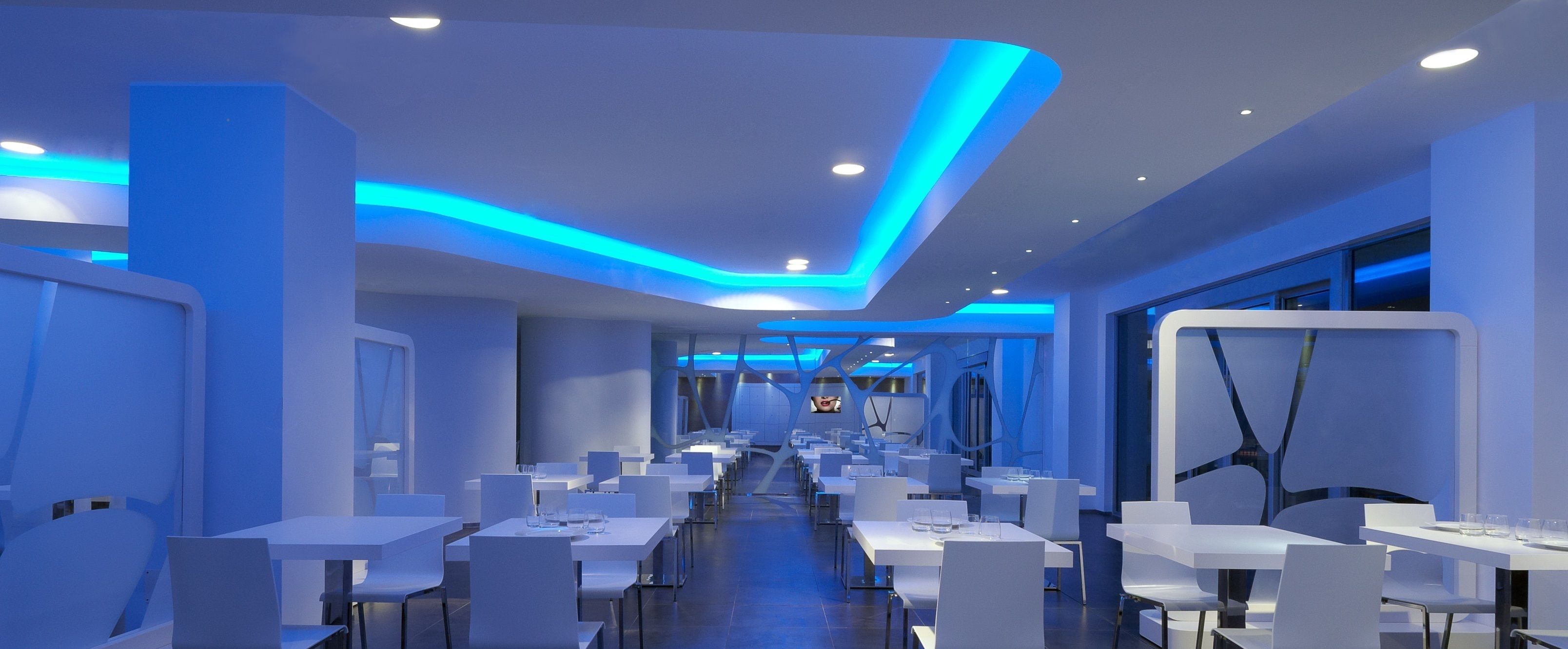 Un espace de restauration moderne avec des tables et des chaises blanches, des éclairages bleus d’accent et un long couloir menant à un comptoir.