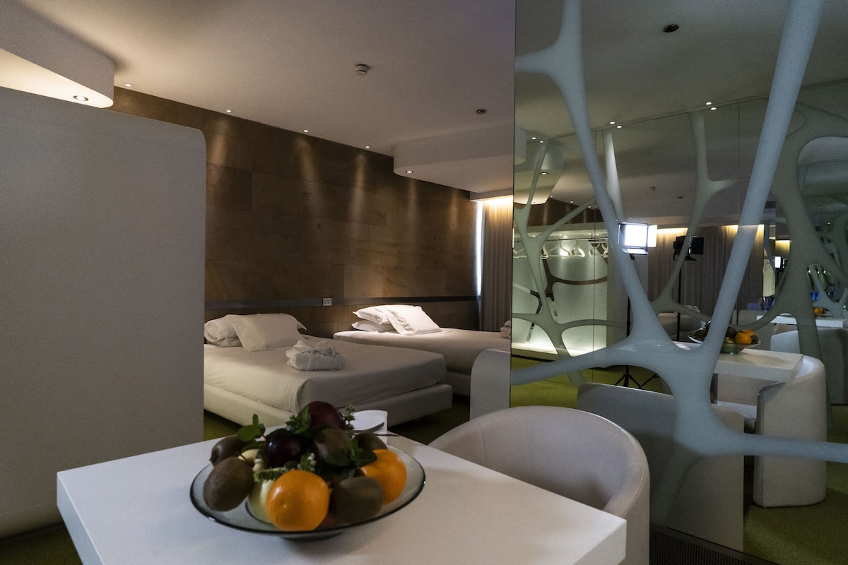 Une chambre d’hôtel moderne avec un lit, une table à manger agrémentée de fruits, et un décor architectural original en arrière-plan.