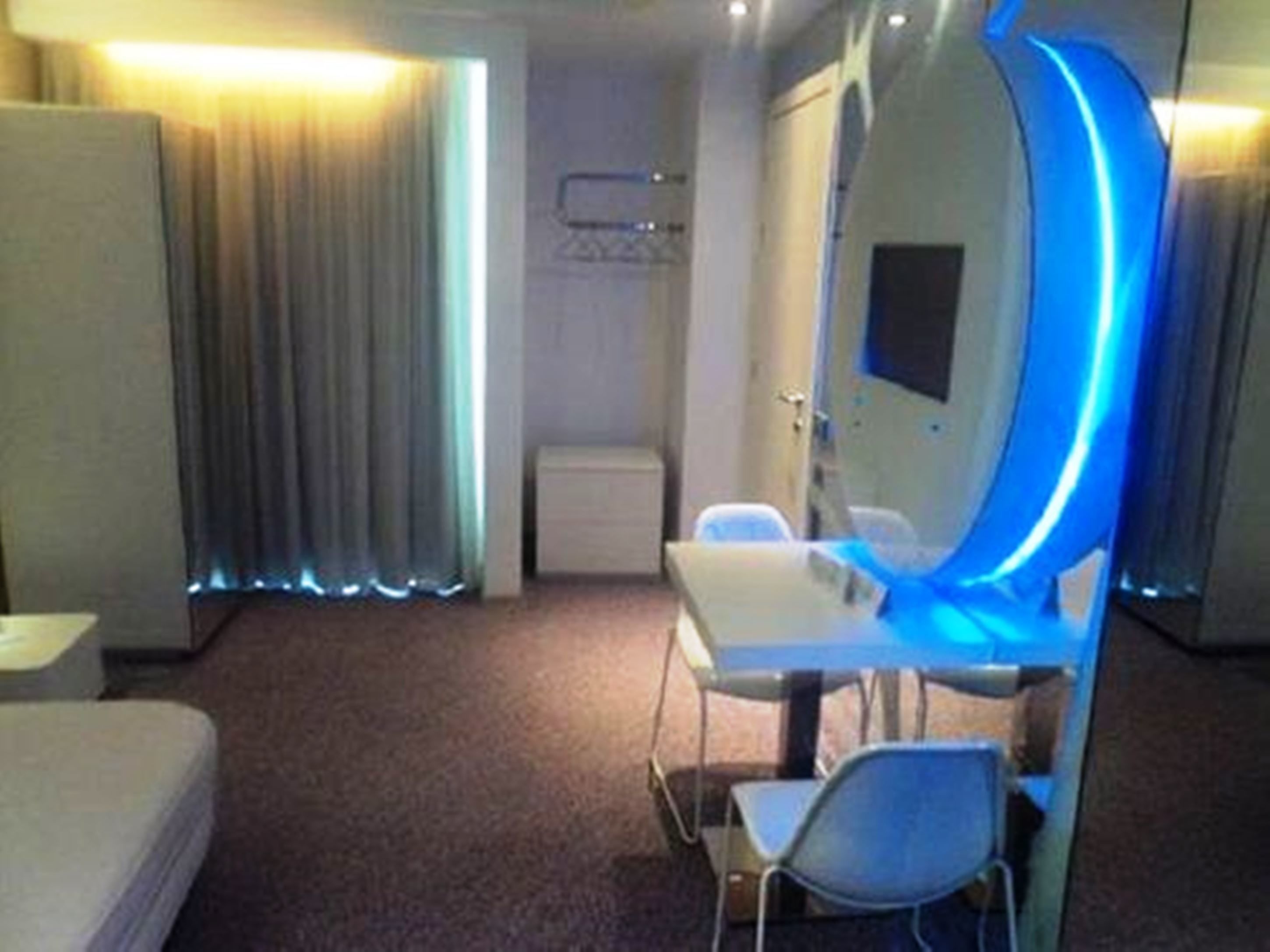 A hotel room with a bed, a desk with a chair, a mirror with blue lighting, and a window with curtains.