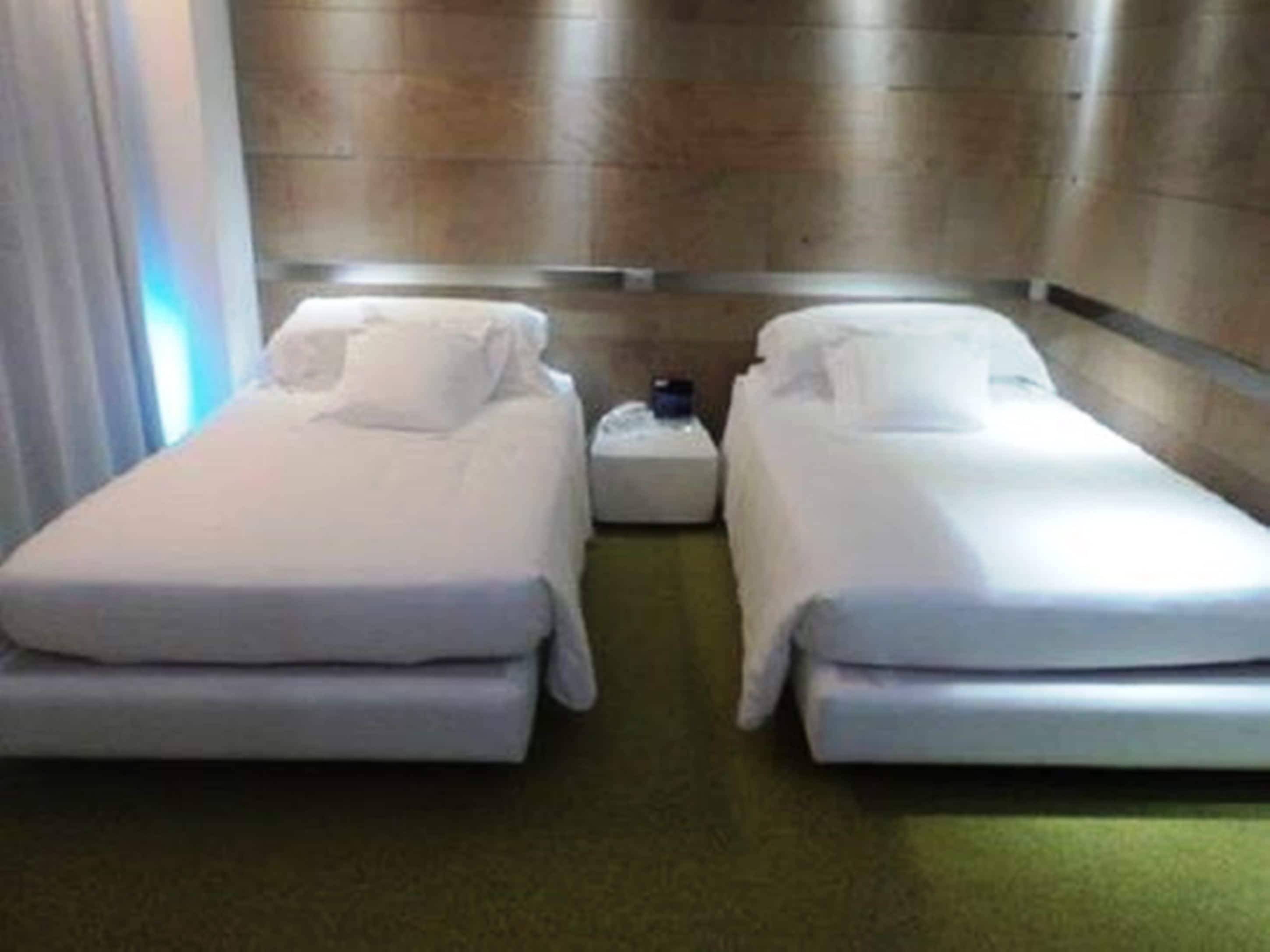 A hotel room with two single beds, a small bedside table, and a light-colored carpet.