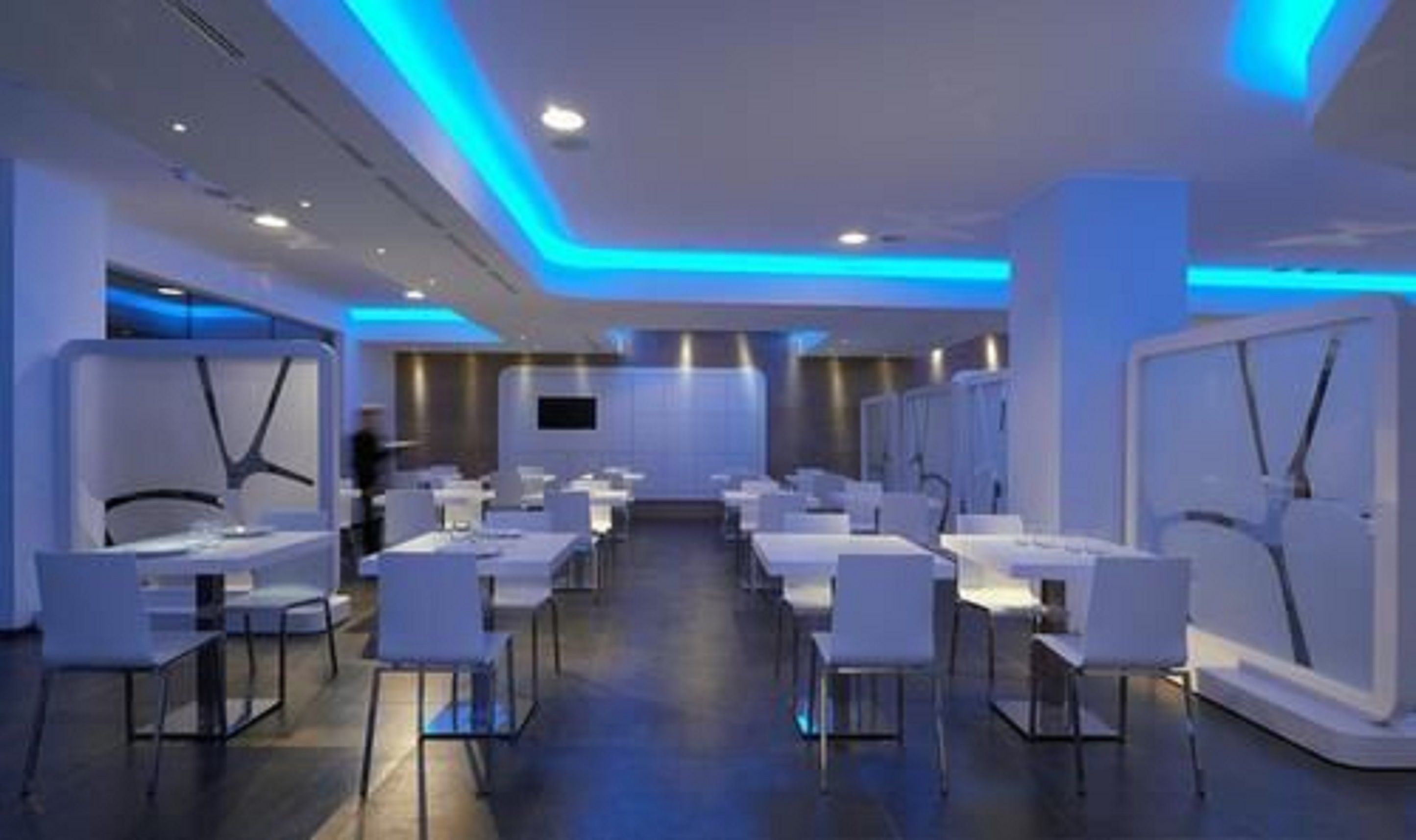Um saguão de hotel moderno, com uma área de recepção central, mobiliário branco e iluminação ambiente azul.