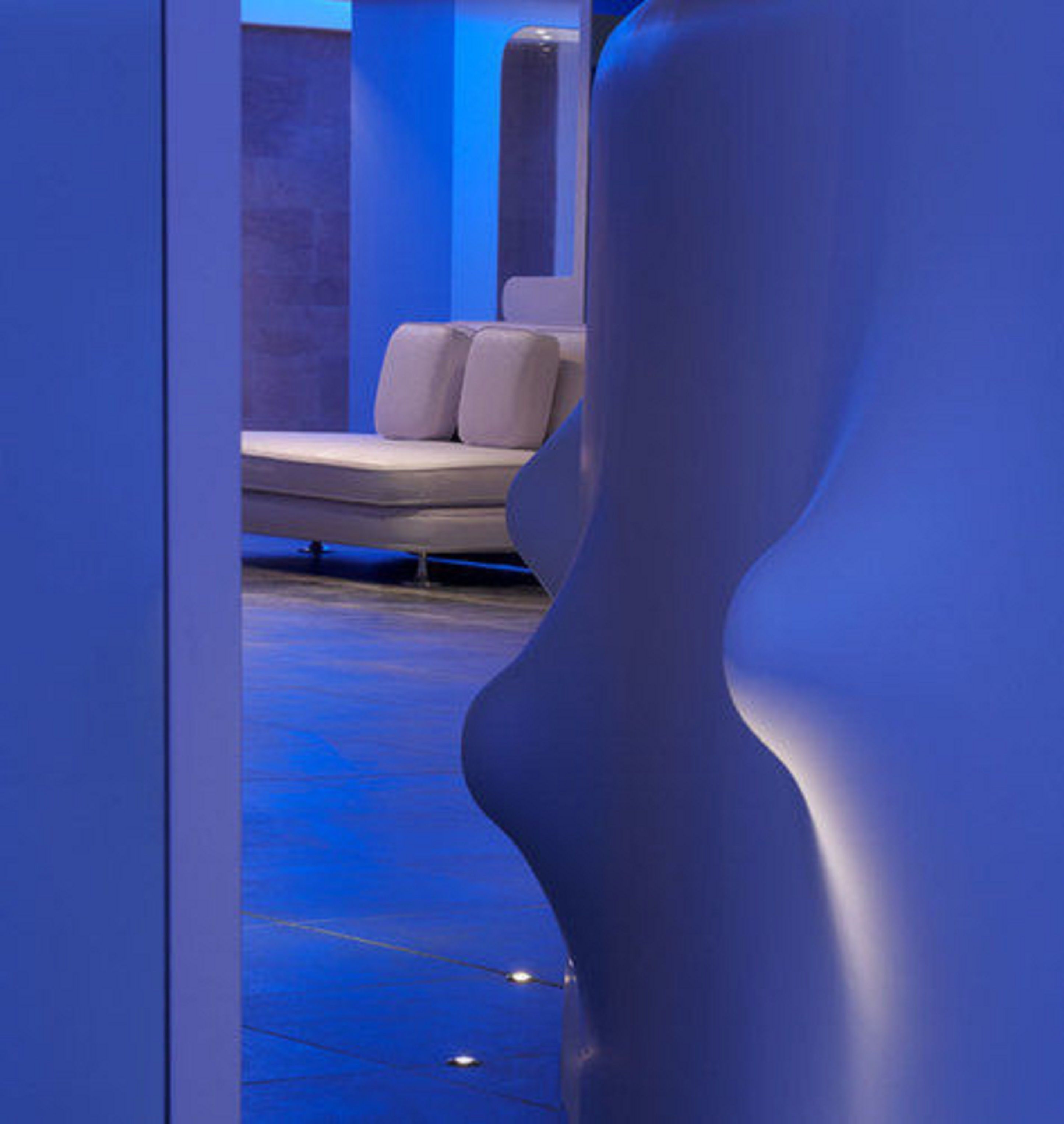 Ein modernes Interieur mit einer gebogenen weißen Struktur, einer Sitzecke mit Sofa und Kissen sowie einer blauen Beleuchtung.
