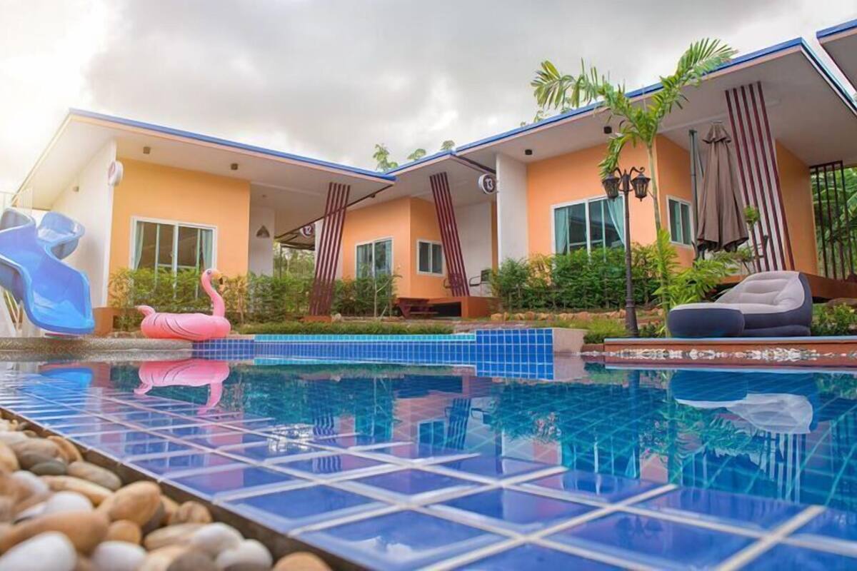 Ein Swimmingpool mit einer rosa Flamingo-Dekoration, einer Liege und einem Gebäude mit blauem Dach und orangenen Wänden.
