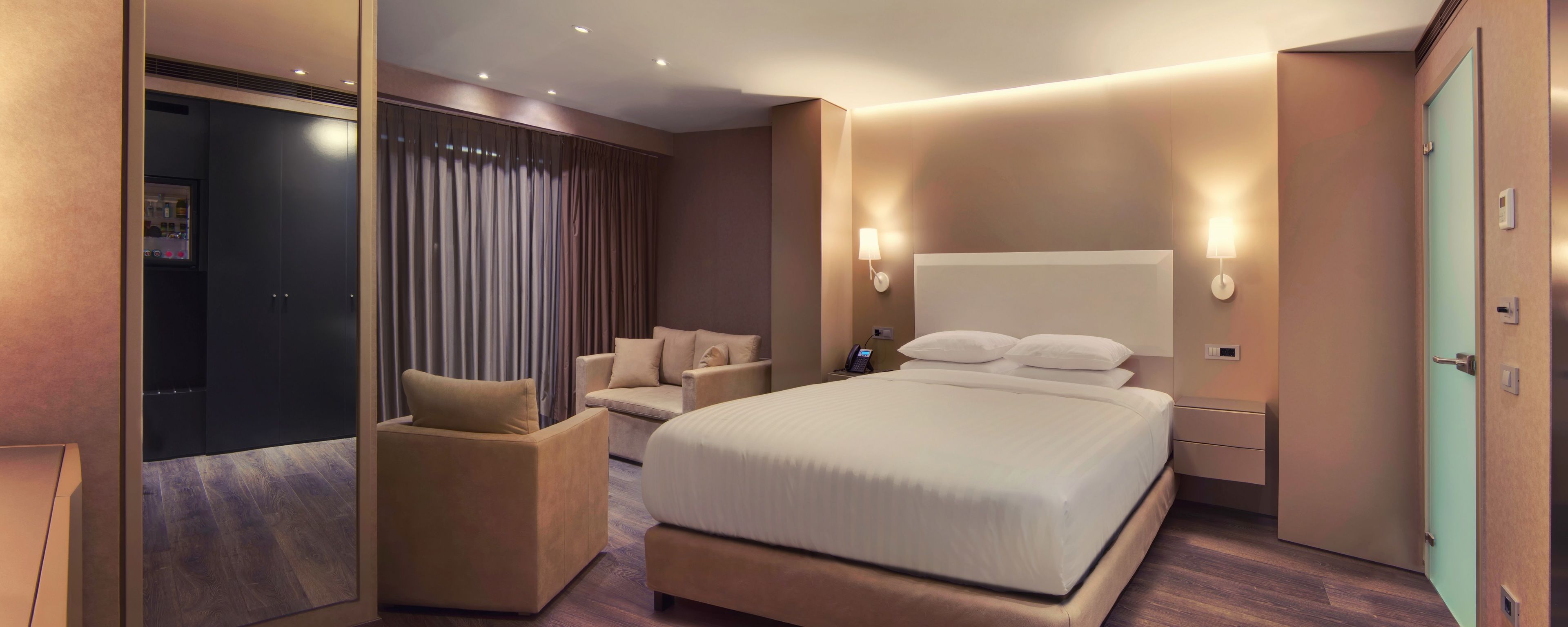 A modern hotel room with a large bed, a sitting area with a sofa, a desk, and a large window with curtains.