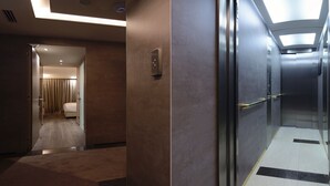 Un couloir d’hôtel avec un ascenseur moderne, une salle de bain visible avec un lit, et un sol recouvert de moquette.