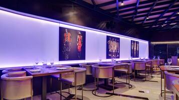 A modern restaurant with high tables and chairs, illuminated by purple lighting, and adorned with large fashion-themed wall art.