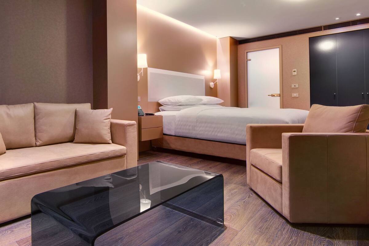 Hotellihuone, jossa on sohva, tuoli, sänky, yöpöytä ja vaatekaappi.